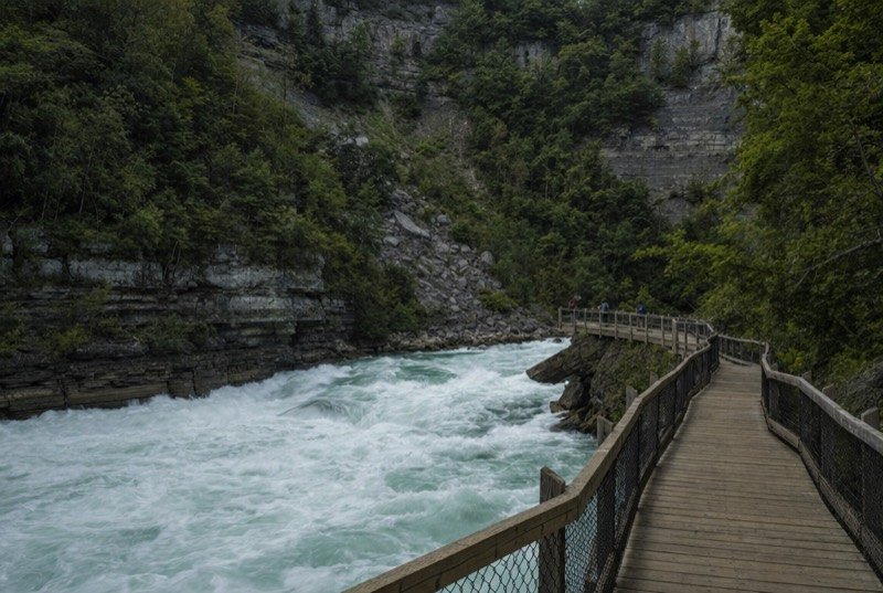 A wooden boardwalk beside white-water rapids at the foot of a steep wooded gorge