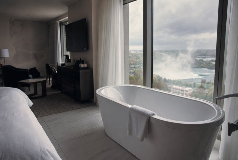 A tidy hotel suite with a deep soaker tub next to a tall window and sheer curtains