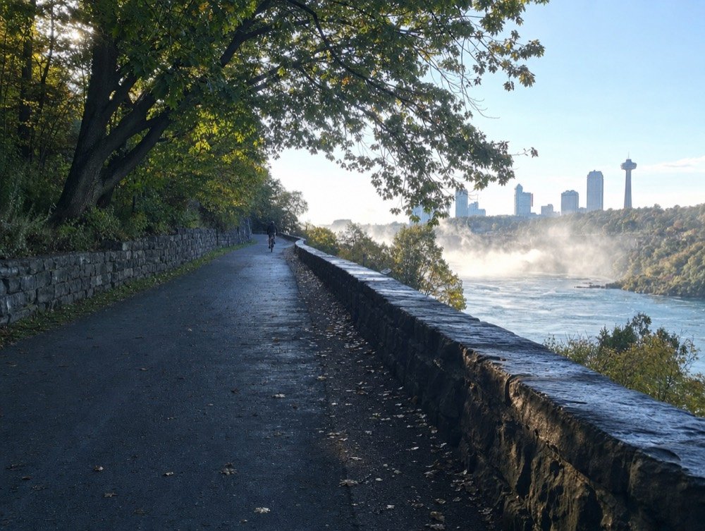 A paved trail beside a riverside parkway with a low stone wall and mature maples, a lone cyclist far ahead in morning light